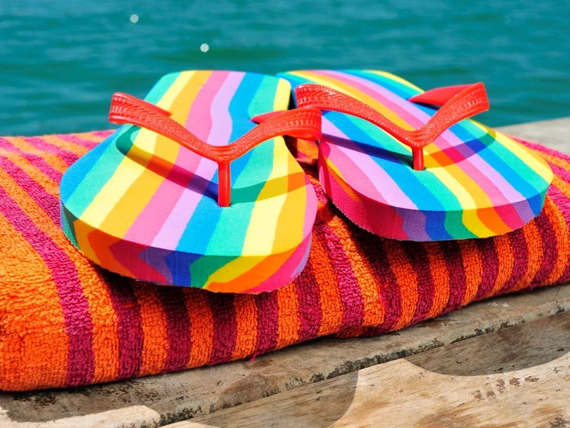 Une paire de tong multicolore est posée sur une serviette de plage pliée sur un ponton en bois avec le lac en fond