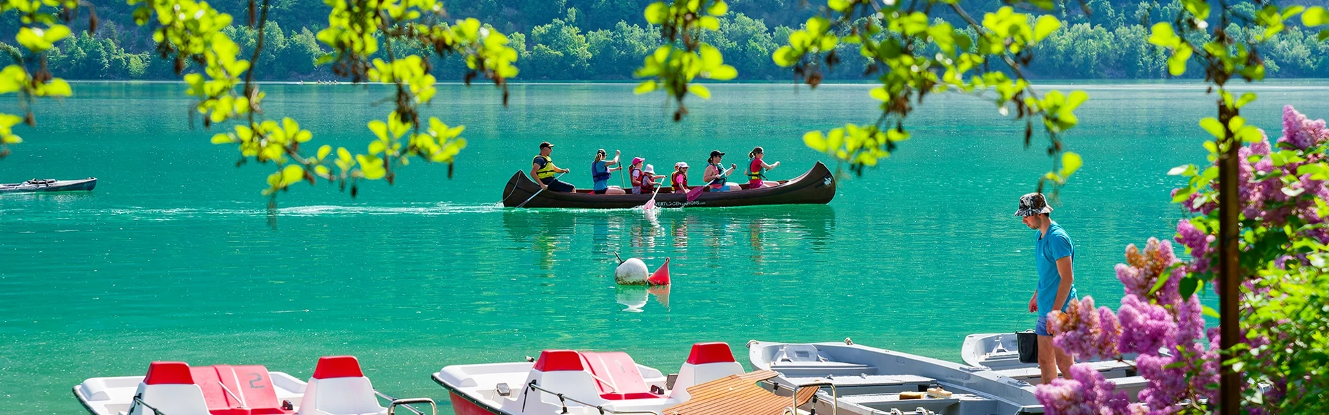 Sur le lac, on voit passer un canoé avec des adultes et des enfants en train de pagayer. Au premier plan des pédalos et bateaux électriques à la location.