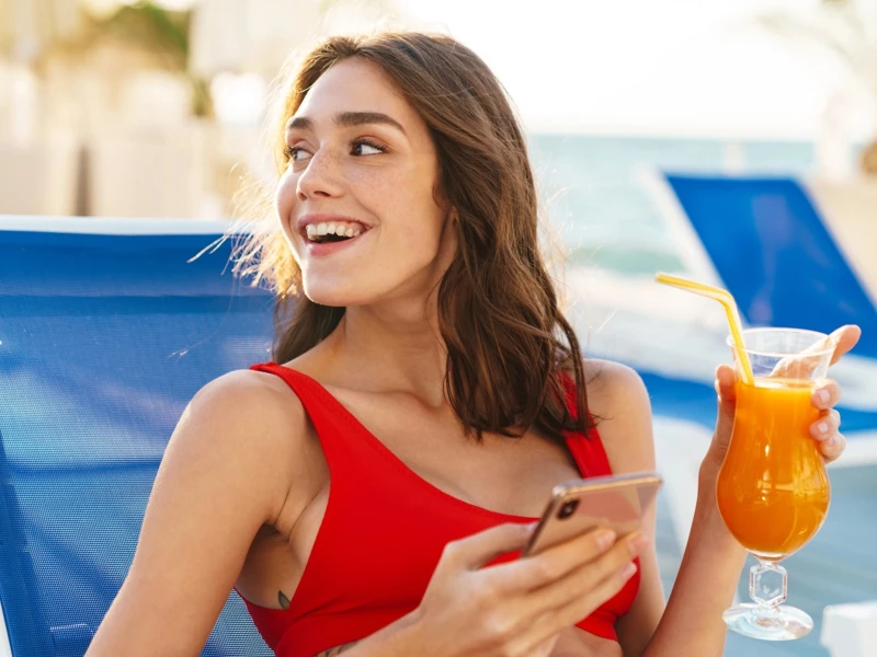 Une jeune femme souriante en maillot de bain rouge est installée sur un transat et tient un cocktail dans la main gauche et son téléphone portable en main droite