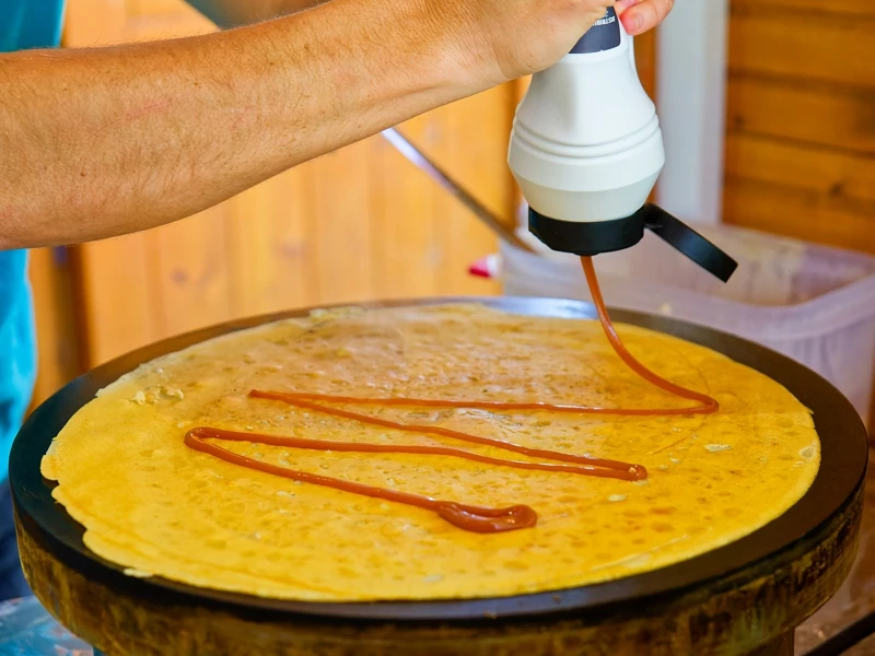 Un homme prépare une crêpe sur une crépière. Il la nappe de caramel.